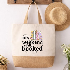 Könnte beinhalten: Eine Canvas-Tragetasche mit beigem Boden und Griffen. Die Tasche zeigt den Text "my weekend is fully booked" mit Illustrationen von Büchern und Sternen. Ein Strohhut hängt über der Tasche.