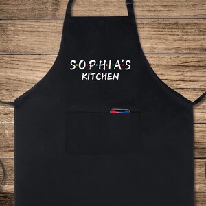 Puede incluir: Delantal de cocina negro con un bolsillo blanco y el texto "SOPHIA'S KITCHEN" en blanco con puntos de colores.