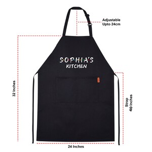 Puede incluir: Delantal de cocina negro con tirantes ajustables. El delantal tiene un bolsillo y dice "Sophia's Kitchen" en texto blanco con puntos de colores.