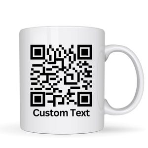 May include: White ceramic mug with a black QR code design and the words "Custom Text" printed below. The mug has a curved handle and a glossy finish, suitable for hot or cold beverages.
