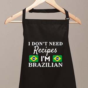 May include: Black apron with the text "I DON'T NEED Recipes I'M BRAZILIAN" in white. The apron features two Brazilian flags. The apron is hanging on a wooden hanger.
