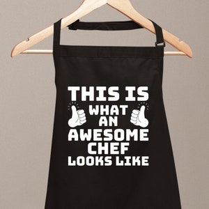 Könnte beinhalten: Schwarze Schürze mit weißem Text, der "Das ist, wie ein großartiger Koch aussieht" lautet, mit zwei Daumen hoch-Symbolen auf beiden Seiten des Textes.