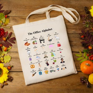 Puede incluir: Una bolsa de tela blanca con una ilustración en blanco y negro del alfabeto de "The Office". Cada letra está representada por un personaje u objeto del programa.