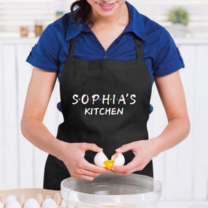 Puede incluir: Un delantal negro con el texto "SOPHIA'S KITCHEN" en blanco. El delantal tiene tirantes ajustables y un bolsillo grande. Una persona lleva el delantal y est&aacute; rompiendo un huevo.