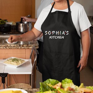 Puede incluir: Delantal de cocina negro con un bolsillo blanco y el texto "Sophia's Kitchen" en blanco con puntos de colores.