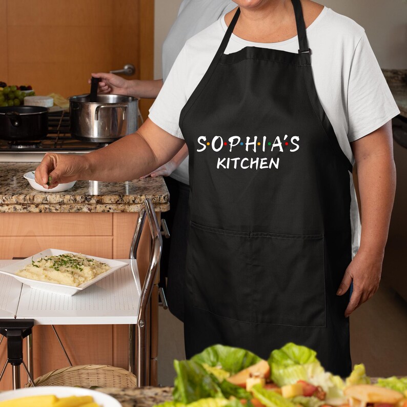 Puede incluir: Delantal de cocina negro con un bolsillo blanco y el texto "Sophia's Kitchen" en blanco con puntos de colores.