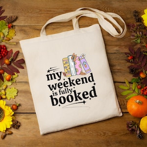 May include: A cream-coloured tote bag with the text "my weekend is fully booked" in black. The bag features a graphic of three colourful books with floral and mushroom designs. The bag has a natural canvas texture and long handles.