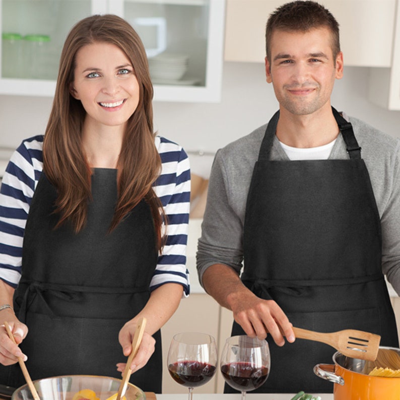 Puede incluir: Dos personas con delantales negros est&aacute;n cocinando en una cocina. Ambos sonr&iacute;en y sostienen utensilios de madera. Hay dos vasos de vino tinto en la encimera.
