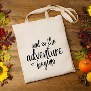 May include: A white canvas tote bag with black text that reads "and so the adventure begins" with a small heart symbol before the word "begins".
