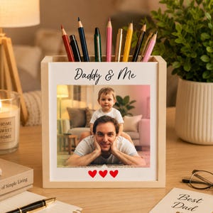 May include: A light-colored wooden pencil holder featuring a printed photograph of a man and a child. The text Daddy & Me is printed above the photo, and three red hearts are printed below. Several colored pencils are stored inside the container.
