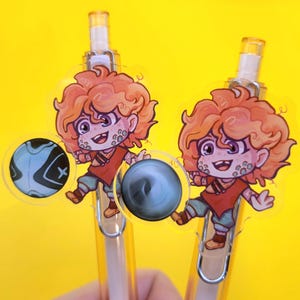 May include: Two clear pens with cartoon character toppers. The toppers feature a red-shirted character with orange hair and a smiling expression. Each pen has a circular design on the side, one blue and black, the other gray.