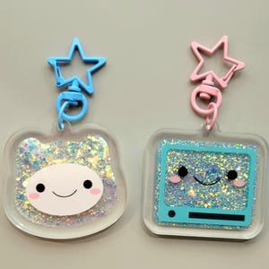 May include: Two glitter keychain charms with star-shaped clips. One charm is a clear square with a blue cartoon TV inside, and the other is a clear oval with a white cartoon face inside. Both charms have iridescent glitter inside.