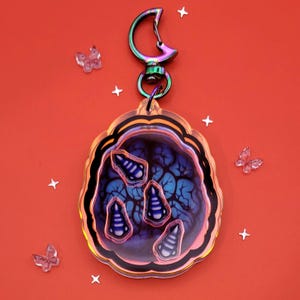 Puede incluir: Un amuleto iridiscente de colores con un clip en forma de luna y un amuleto redondo de acrílico transparente con un diseño azul y morado con cuatro figuras estilizadas en su interior. El amuleto está sobre un fondo rojo.