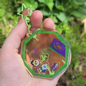 Pode incluir: Um porta-chaves octogonal transparente com uma borda verde e um fecho em forma de estrela verde. No interior, existem várias figuras de personagens de desenhos animados e um elemento arquitetónico roxo e verde. O fundo é de cor laranja quente.