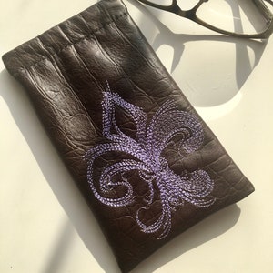 Könnte beinhalten: Eine braune Leder-Brillentasche mit einem gestickten Fleur-de-lis-Muster in Lila.