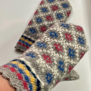 Puede incluir: Un par de manoplas de punto gris con un patrón geométrico de estrellas en blanco, azul y rojo. Los puños presentan rayas horizontales en amarillo, azul y rojo, rematadas con un borde festoneado. La textura del tejido es visible.