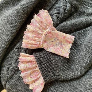 May include: Close-up of a dark grey knitted sweater with decorative pink lace cuffs. The cuffs feature a floral pattern in shades of pink, yellow, and white. The sweater has a textured knit design.