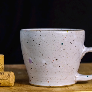 May include: A white ceramic mug with a speckled design and a distinctive handle, with two corks beside it. The mug has colourful speckles and sits on a wooden surface against a black background. The corks have text.