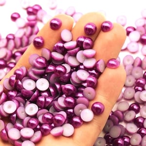 May include: A handful of purple half-round beads with a pearlescent finish. The beads are a deep magenta color and have a flat, white base. The beads are scattered around the hand, with some whole beads visible.