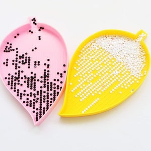 May include: Two leaf-shaped trays for organizing diamond painting beads. One tray is pink and filled with black beads. The other tray is yellow and filled with white beads.