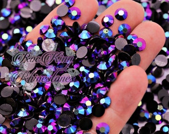 May include: A close-up of a hand holding a large quantity of iridescent purple and black rhinestones. The rhinestones are scattered and have a variety of sizes and shapes.