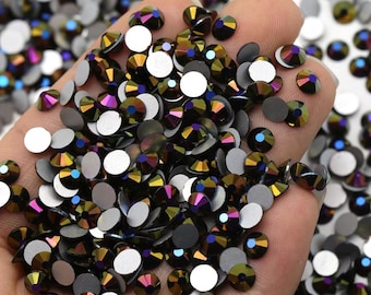 May include: A close-up of a hand holding a large quantity of iridescent black rhinestones. The rhinestones have a rainbow effect and are scattered in a pile.