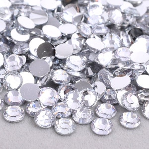 May include: A pile of clear round rhinestones with a silver backing. The rhinestones are scattered on a gray surface.
