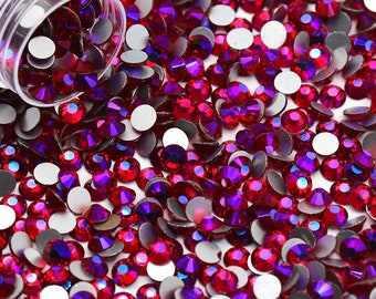 May include: A close-up image of a container of red and purple iridescent rhinestones. The rhinestones are scattered around the container, which is open and spilling out the rhinestones.