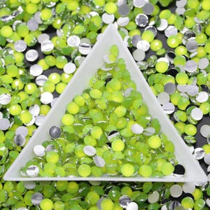 May include: A white triangular tray filled with lime green round rhinestones. The tray is surrounded by more rhinestones on a black surface.