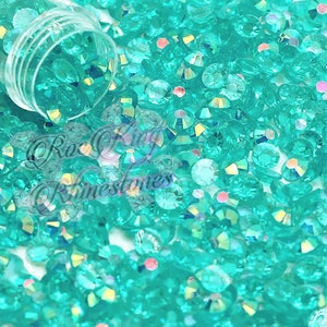 May include: A close-up image of a large quantity of turquoise blue rhinestones. The rhinestones are scattered on a white surface.