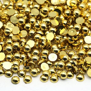 Könnte beinhalten: Ein großer Haufen goldfarbener runder Flachrundcabochons. Die Cabochons sind auf einer weißen Oberfläche verstreut.