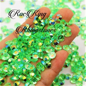 May include: A close-up of a hand holding a large quantity of small, round, green rhinestones. The rhinestones have a faceted, iridescent finish.
