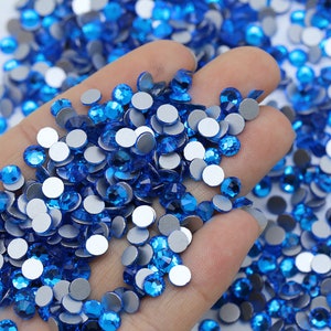 May include: A close-up of a hand holding a large quantity of small, round, blue rhinestones with silver backs. The rhinestones are scattered on a white surface.