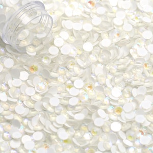 May include: A close-up of a large quantity of white iridescent round rhinestones. The rhinestones are scattered on a white surface and appear to be spilling out of a clear plastic container.