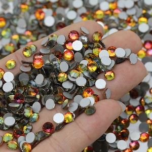 May include: A close-up of a hand holding a large quantity of small, round, iridescent rhinestones. The rhinestones are in shades of orange, yellow, and green, and have a silver backing.