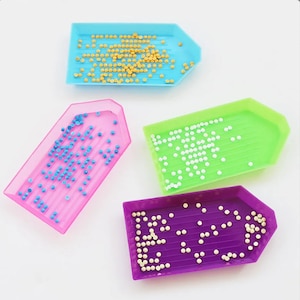 May include: Four diamond painting trays in different colors: blue, pink, green, and purple. Each tray has a pointed end and is filled with small, round beads.