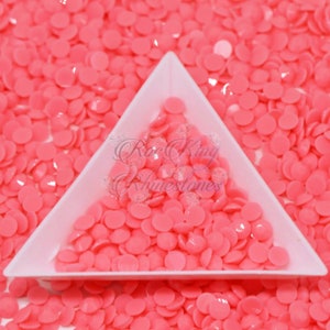 May include: A close-up of a pink, flatback, round, plastic rhinestone. The rhinestones are in a white triangular tray. The rhinestones are scattered around the tray.