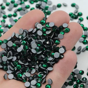 May include: A close-up of a hand holding a large quantity of dark green and silver rhinestones. The rhinestones are small and have a flat back with a silver adhesive.