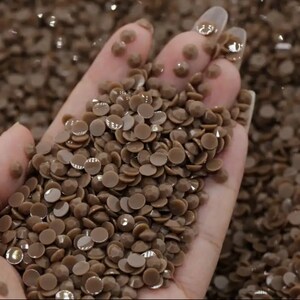 May include: A close-up of a hand holding a pile of small, flat, brown rhinestones. The rhinestones are shiny and have a slightly iridescent finish.