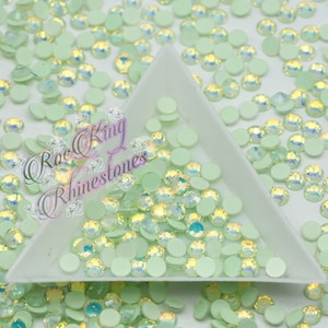 May include: A close-up of a tray filled with light green iridescent rhinestones. The rhinestones are scattered around the tray and some are spilling out of the tray.