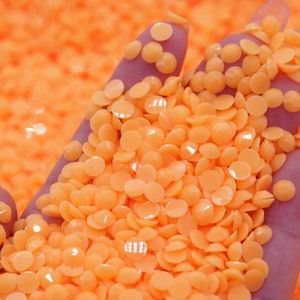 May include: A close-up of a hand holding a large pile of small, round, orange beads. The beads are smooth and shiny, and they are arranged in a random pattern.