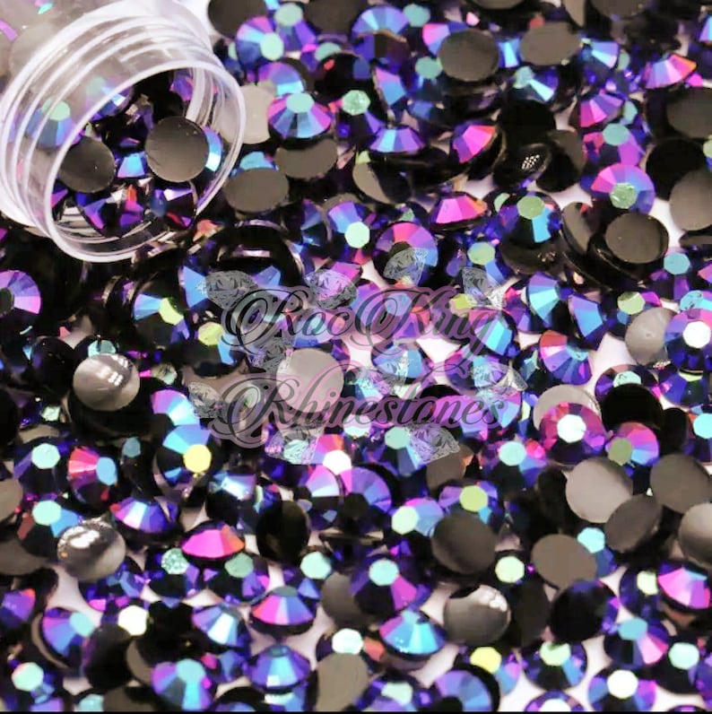 Puede incluir: Una imagen de primer plano de un contenedor lleno de piedras de cristal de color p&uacute;rpura iridiscente. Las piedras de cristal tienen una base negra y un brillo arco&iacute;ris.