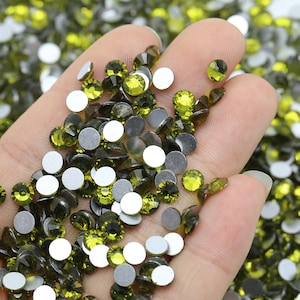 May include: Close-up of olive green rhinestones with silver backing. The faceted crystals are round and reflect light. The rhinestones are scattered, with some held in a hand.