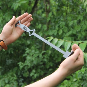 Puede incluir: Una herramienta de metal para asar a la parrilla con una hoja larga y plana y un mango con las iniciales "B.K.". La herramienta se sostiene contra un fondo de follaje verde. El mango tiene un diseño decorativo recortado.