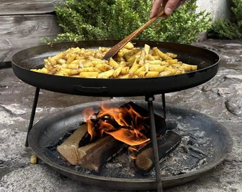 Peut inclure: Une poêle en fonte noire remplie de frites de pommes de terre dorées est en train d'être cuite au-dessus d'un foyer. Le foyer est en métal noir et a une base ronde. Le feu brûle vivement et les flammes lèchent les côtés de la poêle.
