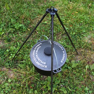 May include: A black cast iron pot with a lid hanging from a tripod stand. The pot has the text "TOTE" and "РОССЛАВЛЯ ВАРЯТСЯ" in Cyrillic script.