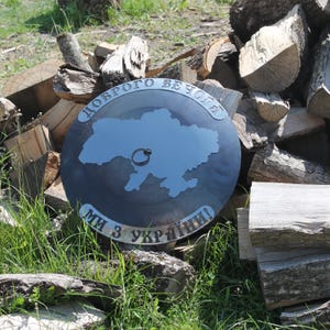 May include: A dark metal decorative piece featuring a map of Ukraine. The piece has the text "Доброго вечора" and "Ми з України!" in a circular design. The piece is set against a backdrop of stacked firewood and green grass.