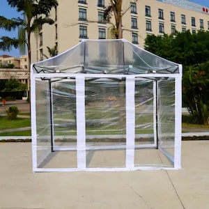 Könnte beinhalten: Ein transparenter, tragbarer Outdoor-Pavillon mit weißem Rahmen und weißen Stoffakzenten. Der Pavillon hat eine rechteckige Form und ist zum Schutz vor Witterungseinflüssen konzipiert. Die transparenten Wände ermöglichen Sichtbarkeit und bieten Schutz.