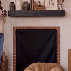 Puede incluir: Una cubierta rectangular negra para chimenea hecha de tela, colocada sobre la abertura de una chimenea de ladrillo. La cubierta presenta un diseño simple y plano con un borde oscuro, sirviendo como pantalla para bloquear el hogar mientras un perro dorado descansa.
