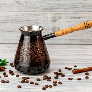 Puede incluir: Una cafetera de metal marrón oscuro con base ancha y cuello estrecho, decorada con intrincados patrones florales en relieve. La cafetera tiene un mango largo de madera clara unido por un conector de metal negro y un borde plateado en la parte superior.